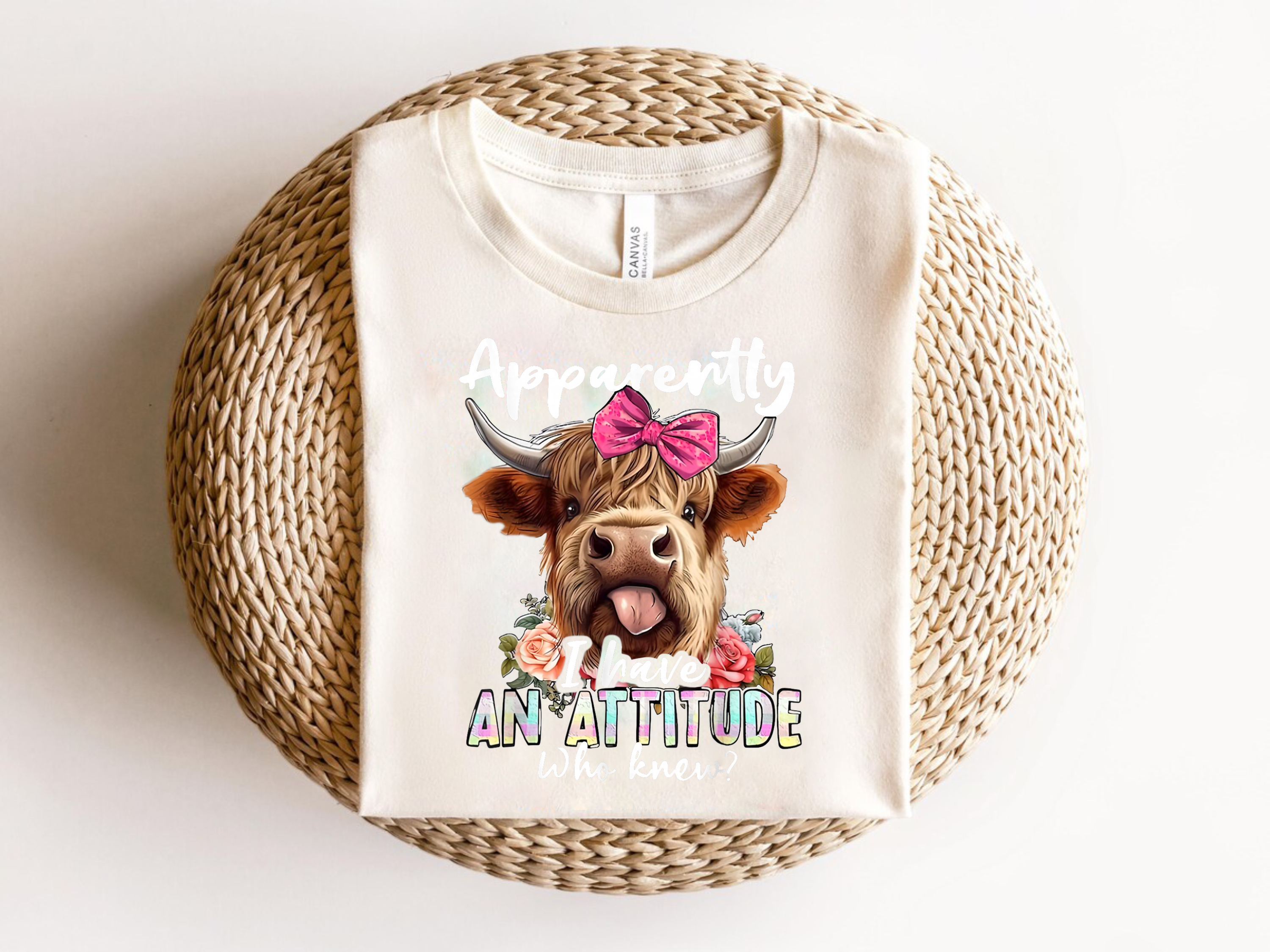 White sweatshirt with a cartoon cow wearing a pink bow and flowers, text 'Apparently I have an attitude', on a woven surface.