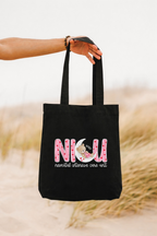 Black tote bag with pink text and graphics held by a person against a blurred natural background