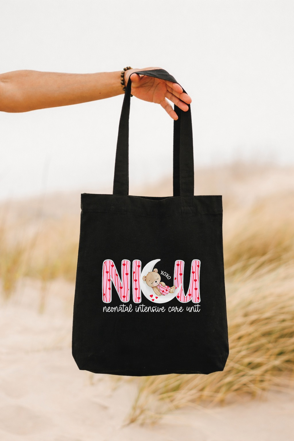 Black tote bag with pink text and graphics held by a person against a blurred natural background
